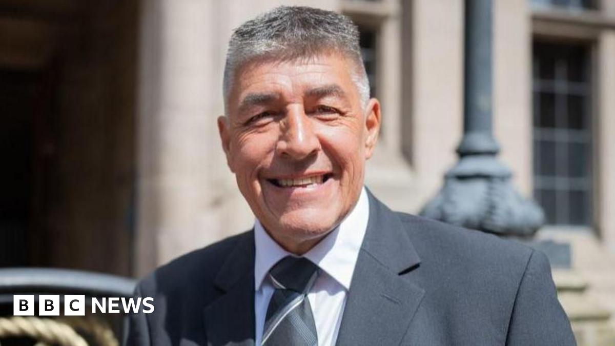 Chris Large is standing in front of the county council building, he is wearing a suit and tie and smiling at the camera.