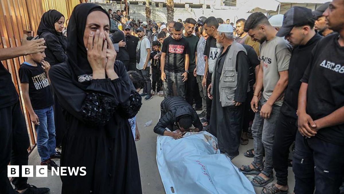 A body bag lies on a ground while a man kneels to kiss it. Others gather round while a woman covers her face and cries. 