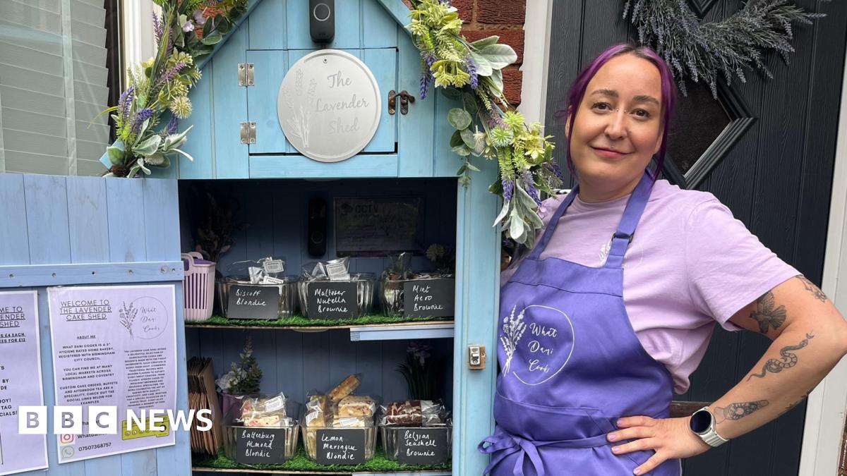 Dani wearing a purple apron and purple t-shirt standing next to the cake shed. The door is open and there are three shelves of cakes. The shed is blue and has a floral garland on the top