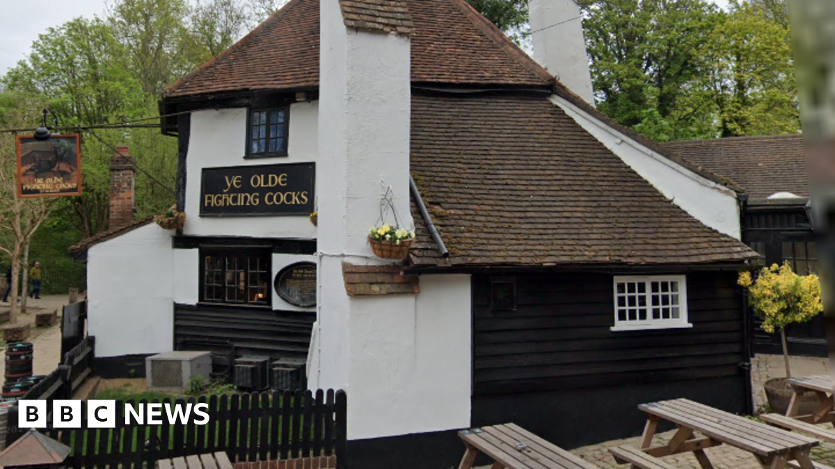 A white building with black painted around the windows. There are two signs with "Ye olde fighting cocks" on them. Trees are in the background.