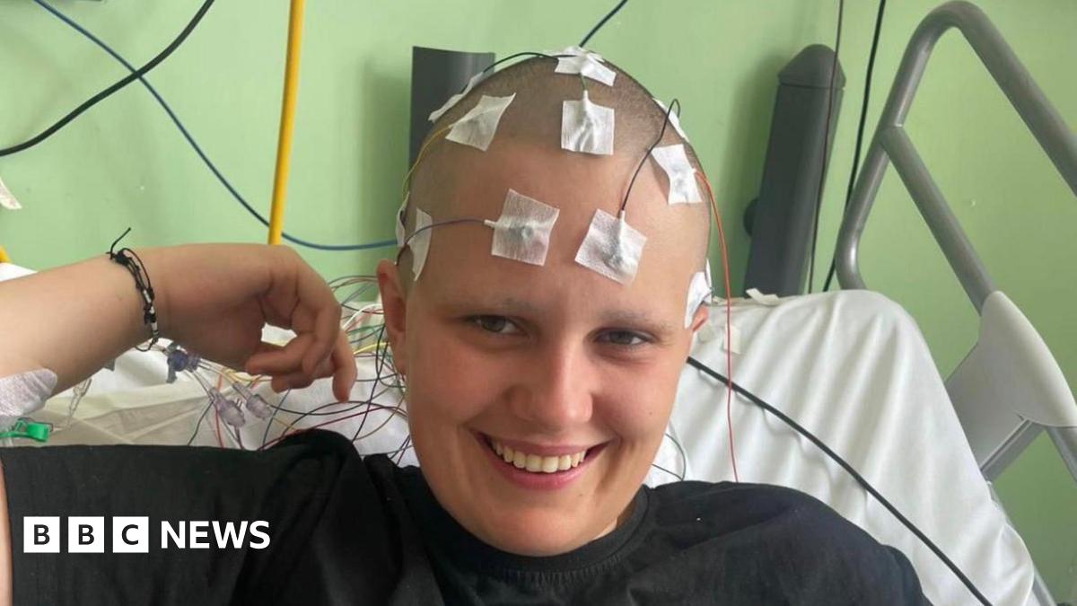 Phoebe is sat in a hosptial bed whilst undergoing a diagnostic test. She is smiling with serval medical wires attached to her head which has been shaved.