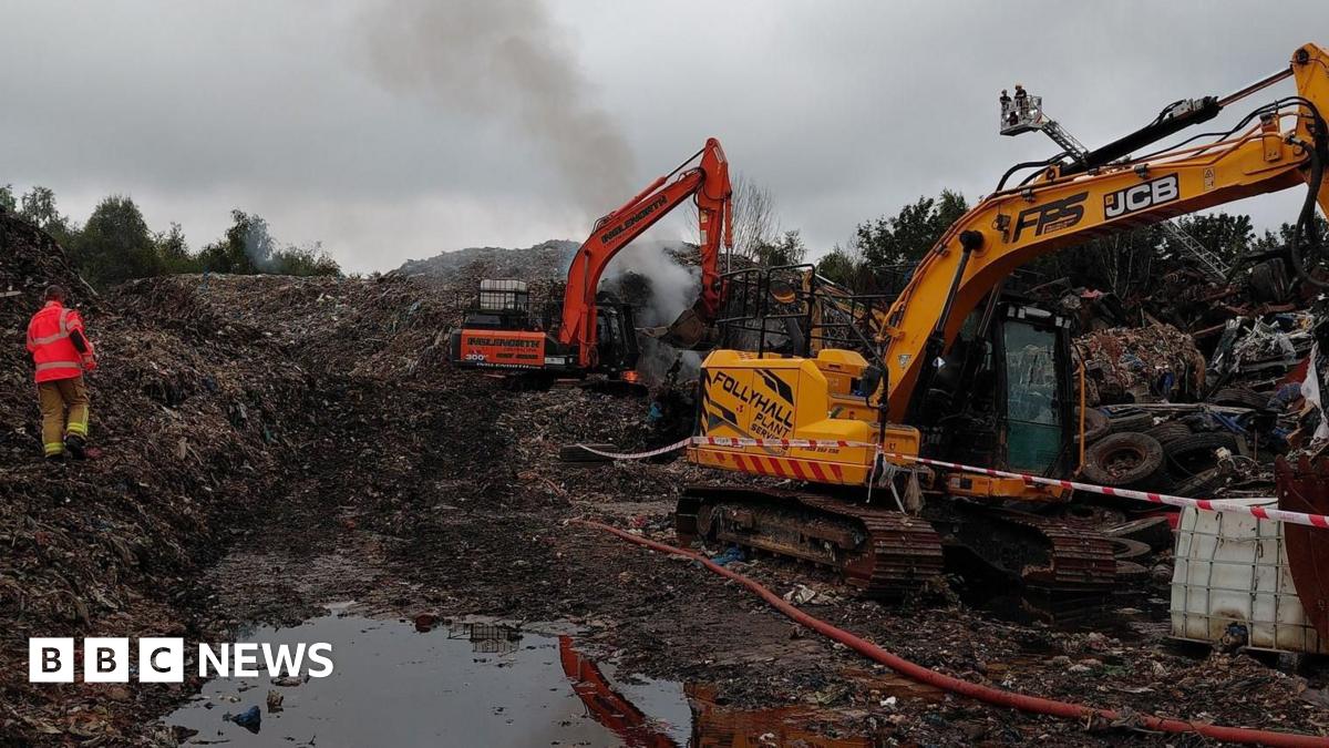 Excavators are parked in the middle of a large waste site. A man in a high vis jacket can be seen walking through it.

