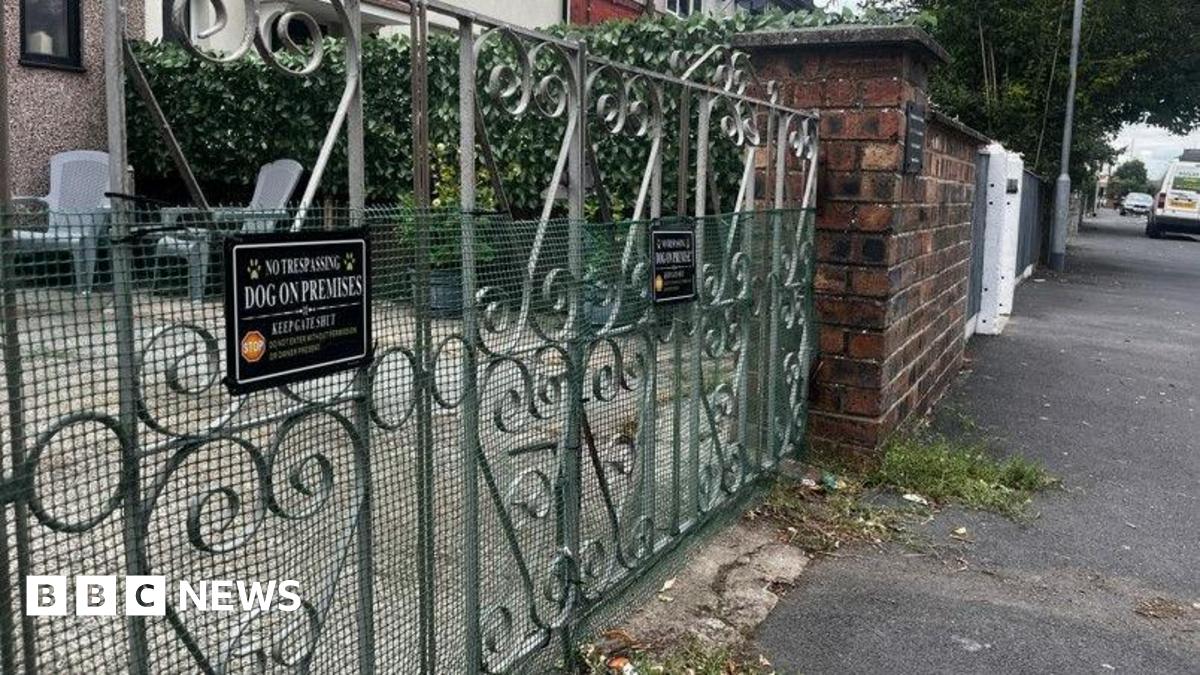 A set of wrought iron garden gates partically covered in a green wire mesh and fixed with a black sign saying 'No Trespassing, Dog on Premises, Keep Gate Shut'. 