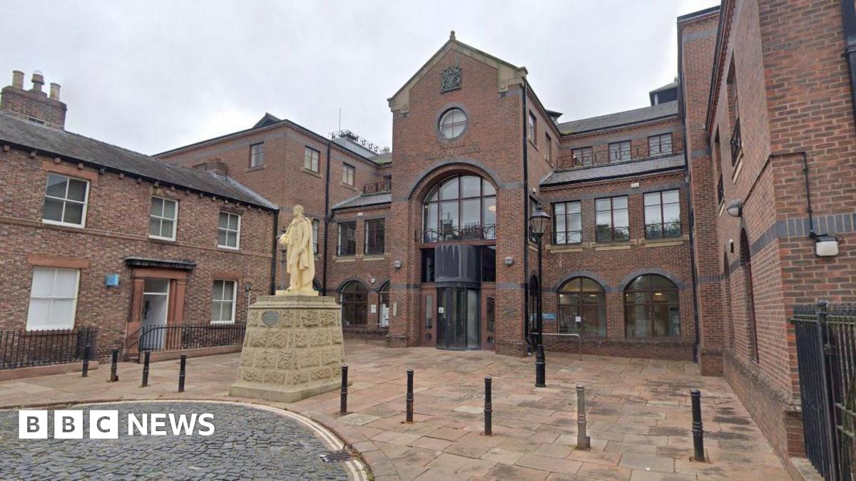 A view of the front of the Carlisle Crown Court building. There is a gold statue on a stone plinth in the centre of a courtyard with thin bollards stretching out in a line either side. The three-storey court building is made with modern dark bricks wit...