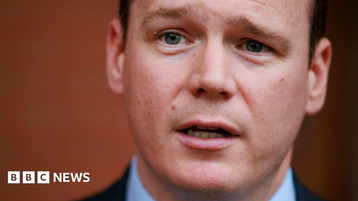 A close up of the Communities Minister Gordon Lyons. He is wearing a blue shirt and dark jacket. He looks like he is in the middle of speaking. 