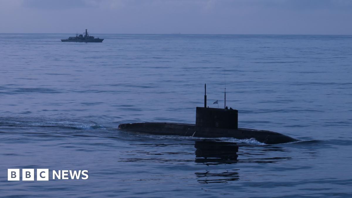 A submarine is seen in the water in the foreground, with a navy warship in the background