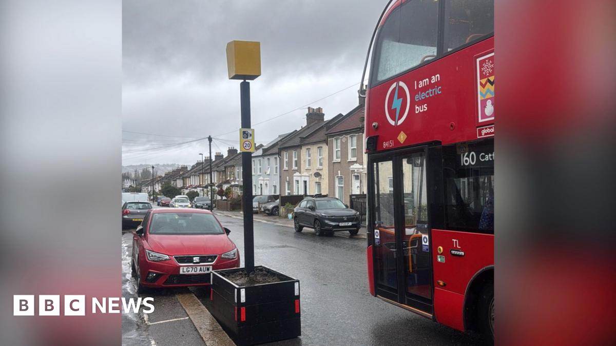 Fake yellow speed camera in a planter next to red double decker 160 bus in the foreground. Parked cars and houses can be seen in the background