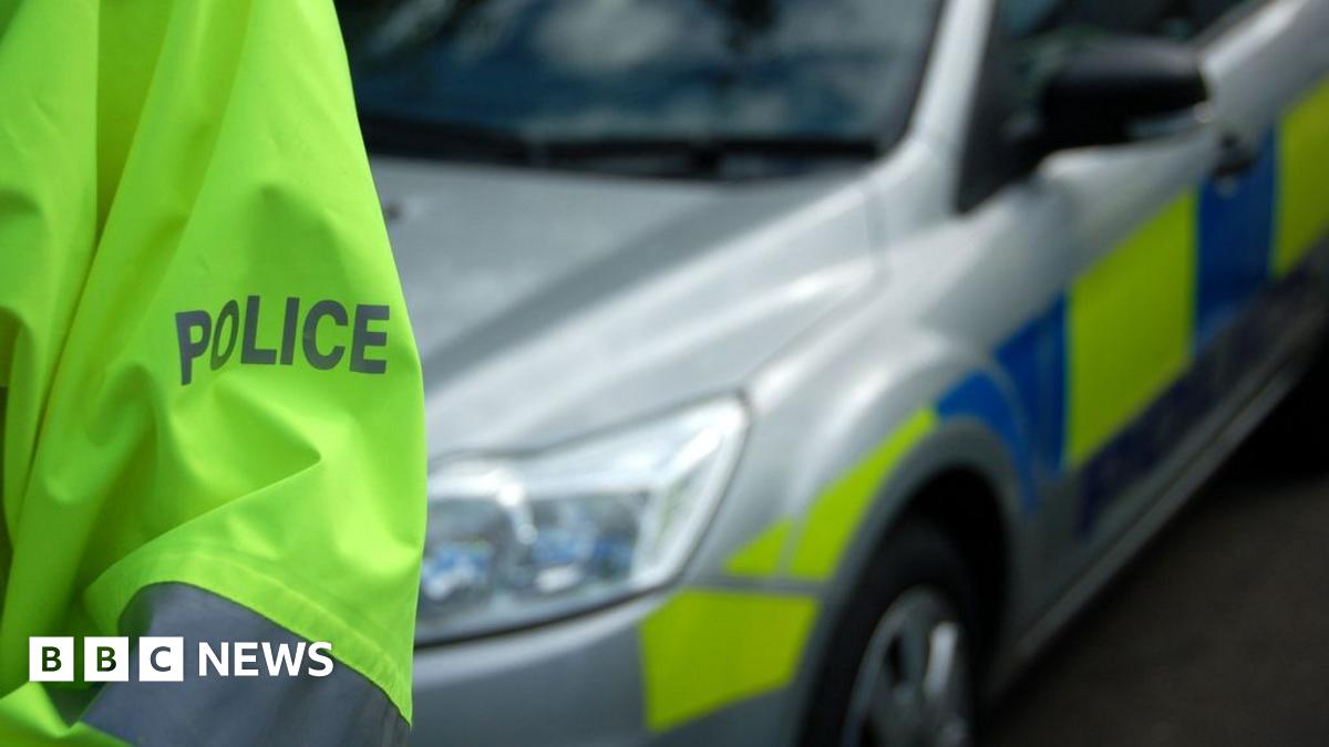 A police car with a police officer in the foreground. All you can see of the officer is the sleeve of his hi-vis jacket which says POLICE