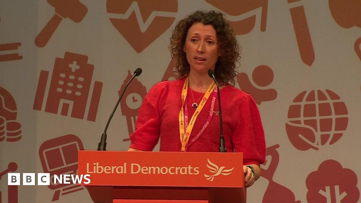 Head and shoulders shot of Dr Zoe Hollowood speaking at Liberal Democrat conference. She has light brown, shoulder-length, curly hair worn loose. She's wearing a puff-sleeved dark orange jumper and stands behind a Lib Dem branded podium.