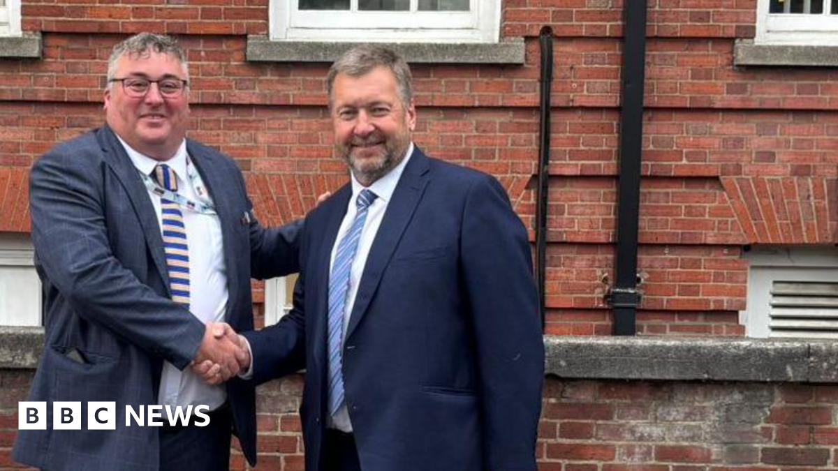 Two men wearing blue suits, white shirts and ties shake hands and smile as they stand outside a red-brick building.