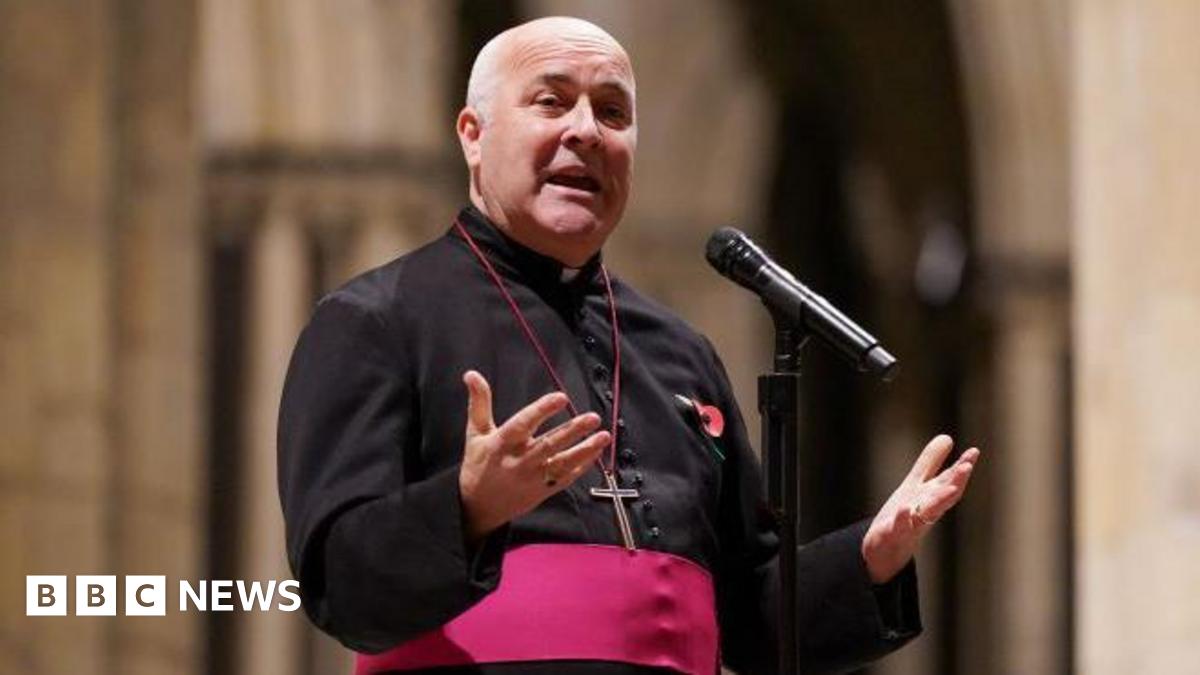 Archbishop of York standing and holding his hands open, palms upwards, wearing his black cassock with red trim