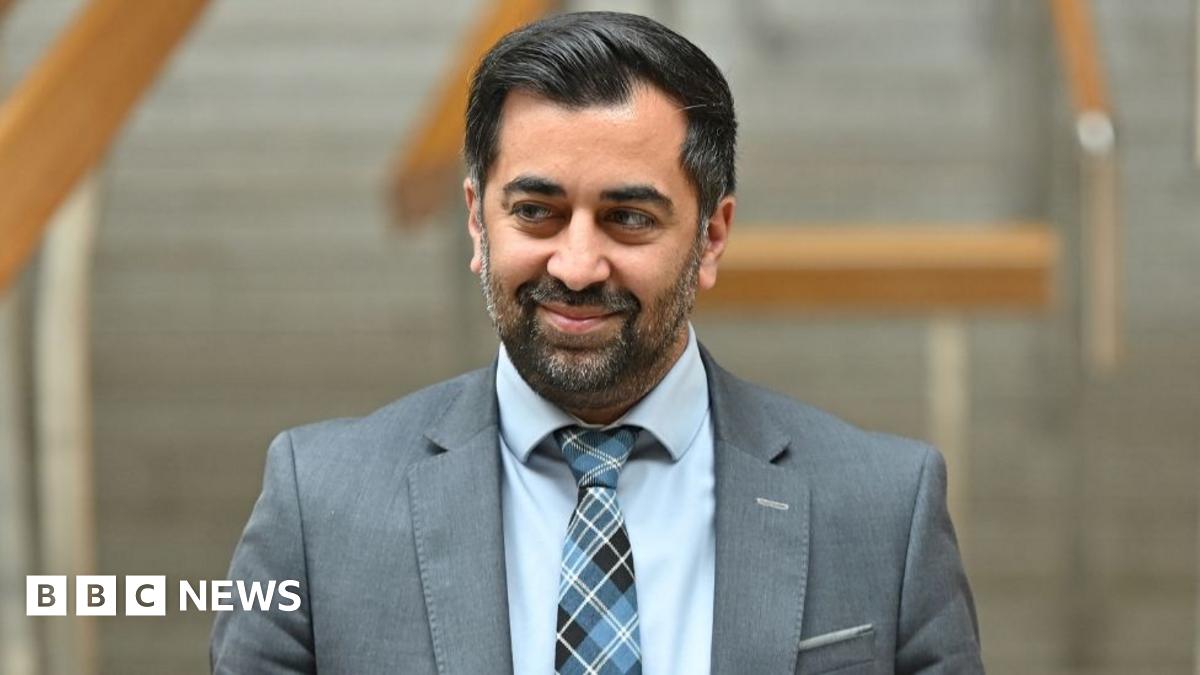 Humza Yousaf, with dark hair and wearing a grey jacket, blue shirt and tartan blue tie, smiles as he looks to his right in a medium close-up shot in the Scottish Parliament 