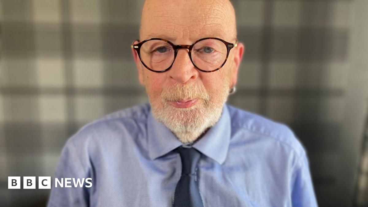 Stuart Hunter is photographed looking directly at the camera. He is a bald man with large round-rimmed glasses and a white beard. He is wearing a blue shirt and dark blue tie.