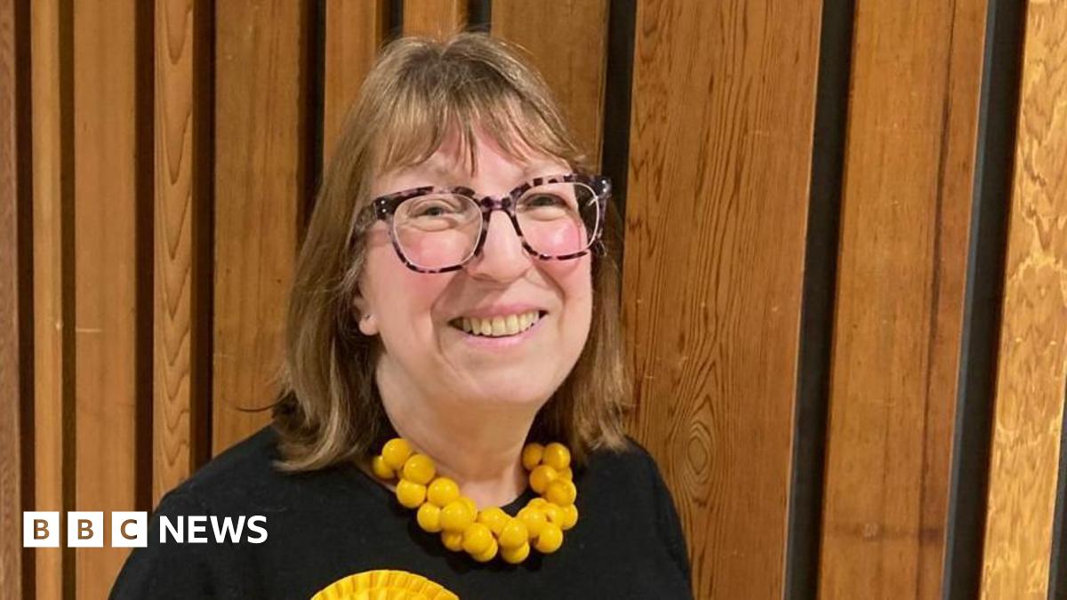 Alison Barnes is smiling at the camera. She has mid-length brown hair and large brown glasses. She is wearing a chunky yellow necklace and a yellow rosette.
