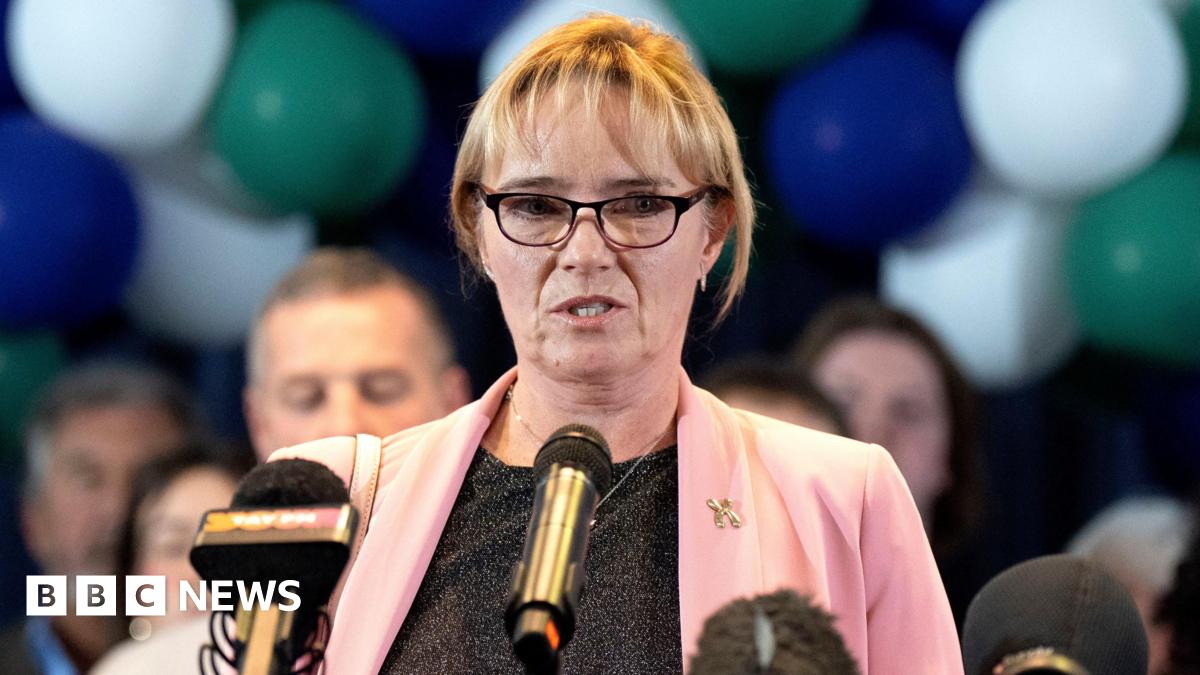 Sandie Peggie in pink jacket and grey blouse, wearing glasses, in front of a mic. There are people behind her.