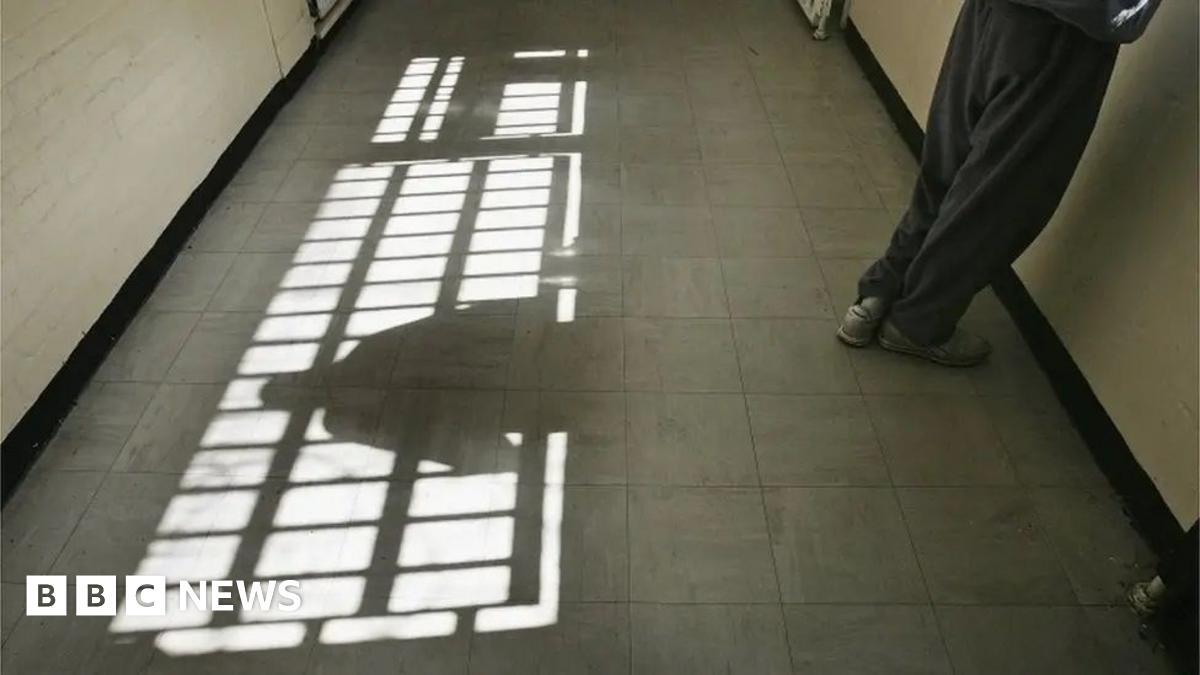 A corridor with sunlight streaming through a barred window, casting shadows on the tiled floor. A person stands against the right wall, only their lower body visible. Their shadow on the floor.