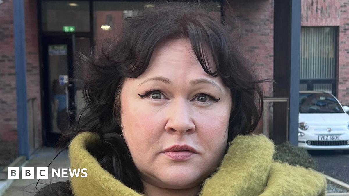 Sara Morrison is wearing a brown top with a large green scarf around her neck. She is standing in front of a red brick building with black windows. There are cars and a motorbike in a car park between her and the building. The path around the building...