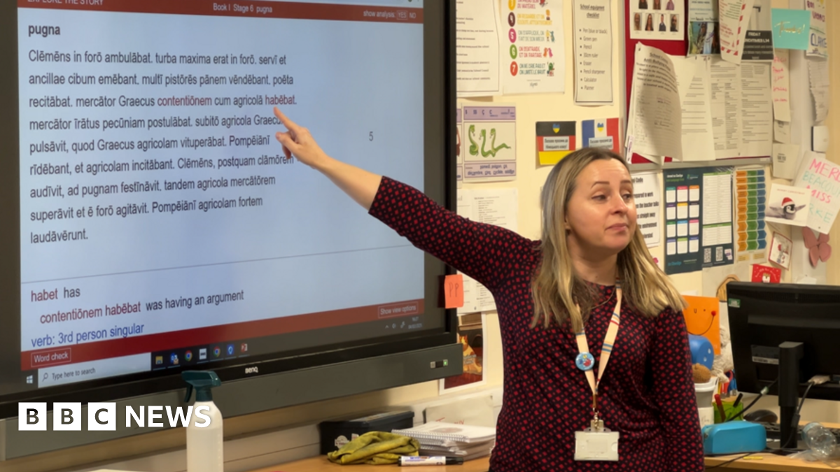 Teacher Marcella Jackson in a classroom. She is pointing behind her to a large TV screen containing Latin words. She has long blonde hair and wears a three-quarter length black top covered in a red spotted pattern and is looking out at the class. She w...