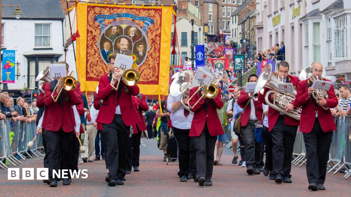 The Durham Miners' Gala. A brass band is walking down the street in red blazers. A red, yellow and blue miners' banner with five men depicted in the middle is being held up behind them. More banners and people can be seen walking behind them with peopl...