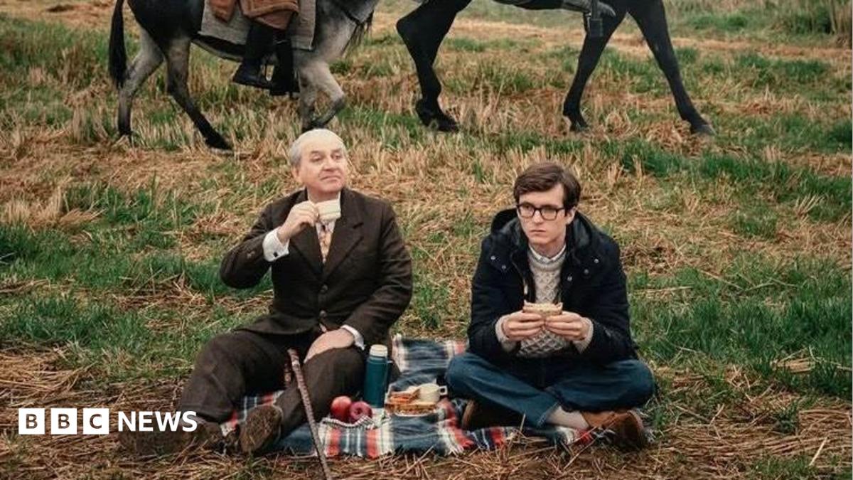 Photo: bbc.co.uk Luis Gnecco in brown suit and Fionn Whitehead in dark duffel coat are having a picnic, sitting on a rug in a field. The