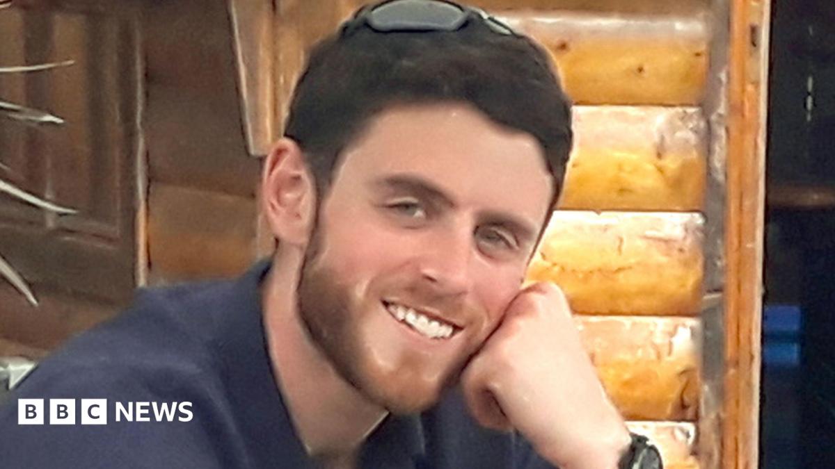 PC Andrew Harper smiles into the camera with his chin resting on his closed left hand. He is wearing a navy top and has a pair of sunglasses perched on the top of his head. He has dark brown short hair and a brown short beard