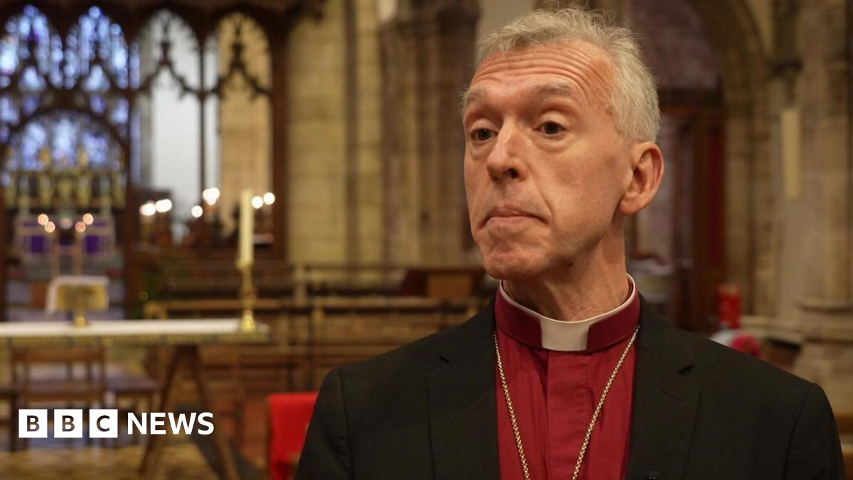 Andrew John in a red shirt and black blazer with a dog collar, looking at the camera in front of an altar. He has grey hair and brown eyes