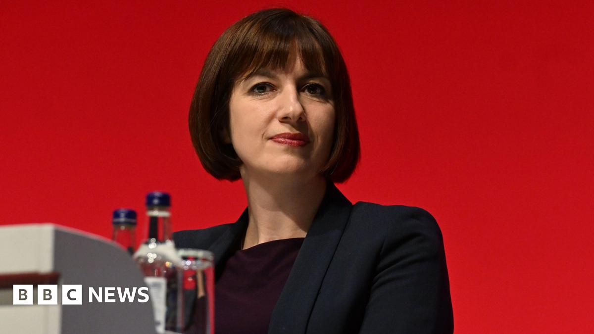 Education secretary Bridget Phillipson, who has short brown hair and a fringe, wears a black blazer and dark top and is pictured against a red background. Bottles and a glass of water are seen in the foreground.