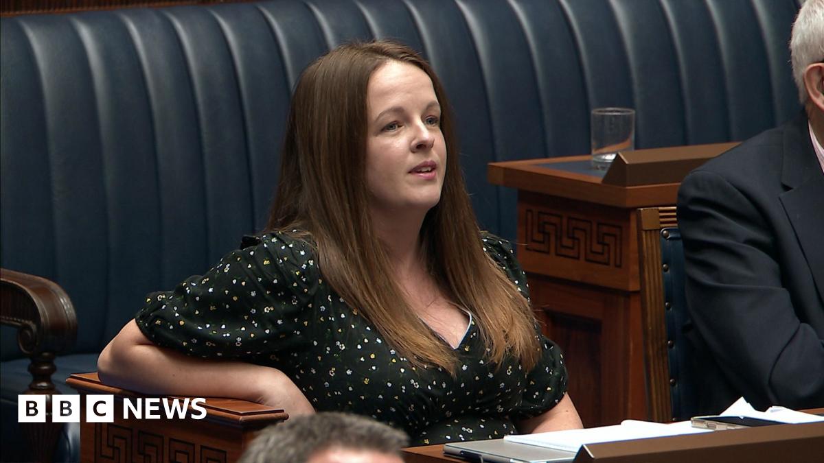 Nuala McAllister, she has long brown hair and is wearing a black floral top, stood in the NI Assembly chamber, there are blue leather seats and wooden tables
