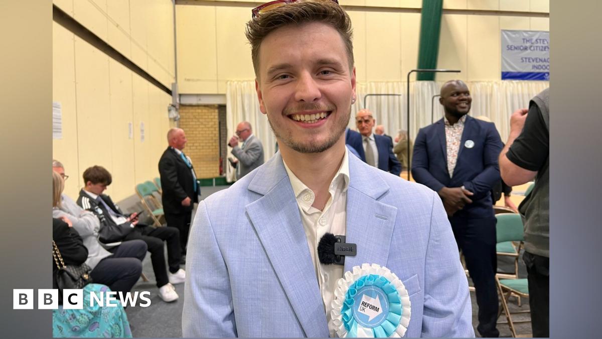 Liam Morrell Phillips smiles at the camera wearing his Reform white and blue badge on a pale blue suit. He has sunglasses pushed back on his head. Other people stand around behind him. 