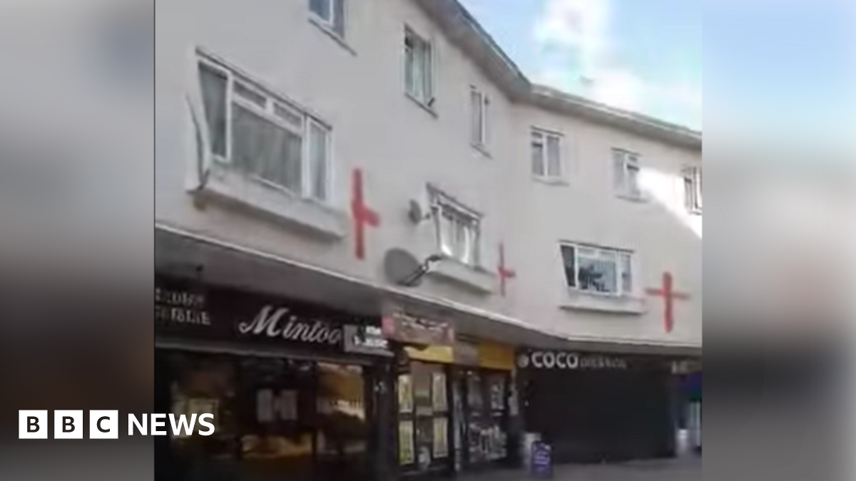 A row of shops is shown on the ground level with flats spread across the two floors above them, with the red flag of St George painted on the white walls of the homes.