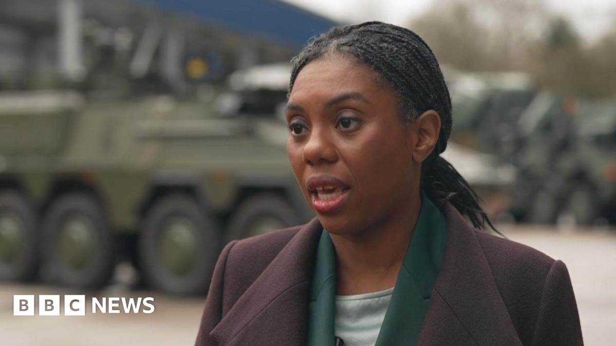 Kemi Badenoch wearing a purple coat, standing in front of a tank. 