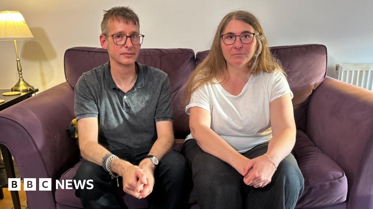 Chris, a man with glasses and wearing a grey polo shirt, is sat on a purple sofa next to his wife Diana, who had long light brown hair and is wearing glasses and a white T-shirt. They are both looking at the camera