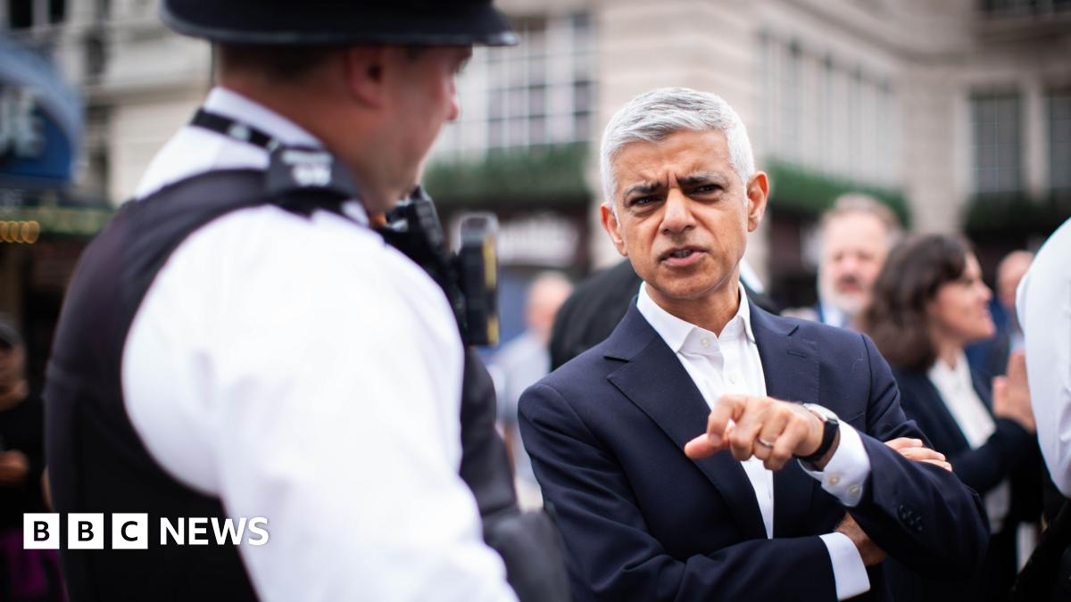 London Mayor Sadiq Khan, wearing a dark suit and white shirt, gestures while speaking to a uniformed police officer in the street, with several people blurred in the background.