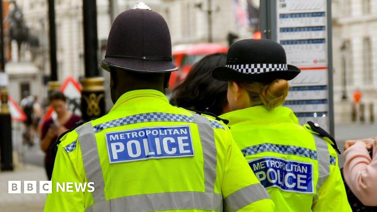 Two police officers, a man and a woman, seen from behind in a busy street walking past a bus stop. Their uniform reads METROPOLITAN POLICE. 