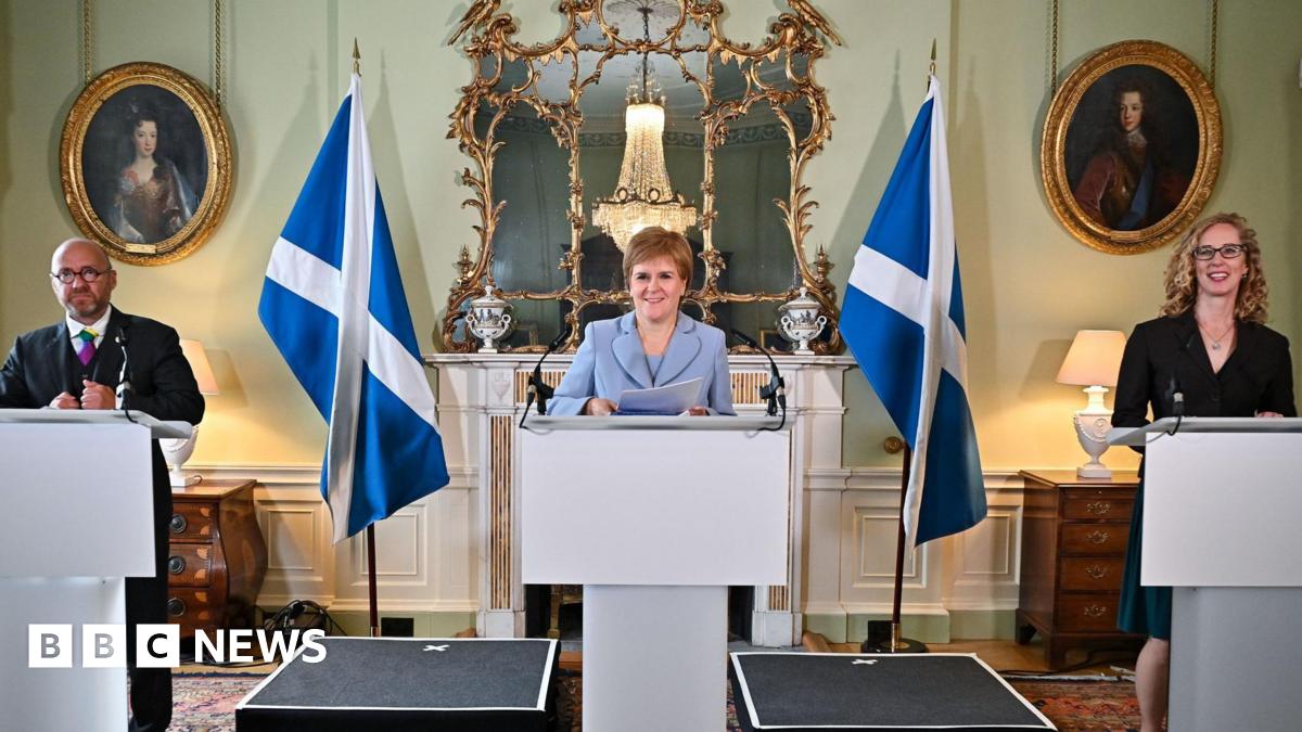 A bald man in a dark suit stood behind a rostrum and to his left is a woman in pale blue suit stood behind a rostrum, and to her left is a woman in a dark dress stood behind a rostrum