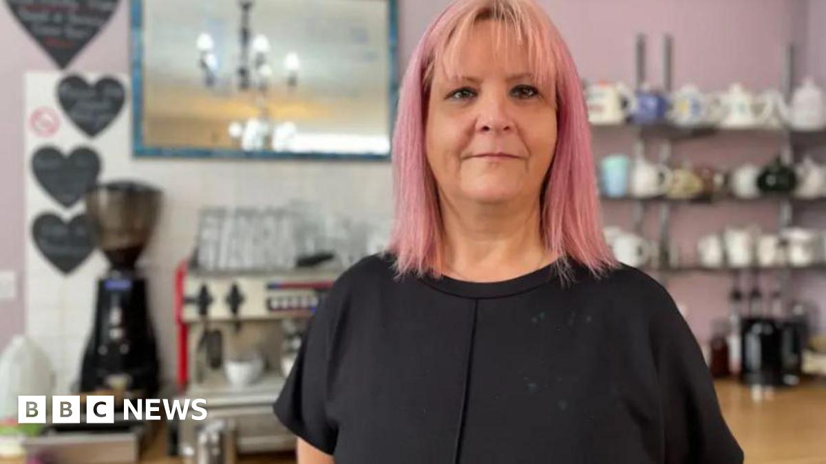 Donna behind the counter of her cafe which has an array of mugs and a coffee machine. She is wearing a black shirt and has pink hair.