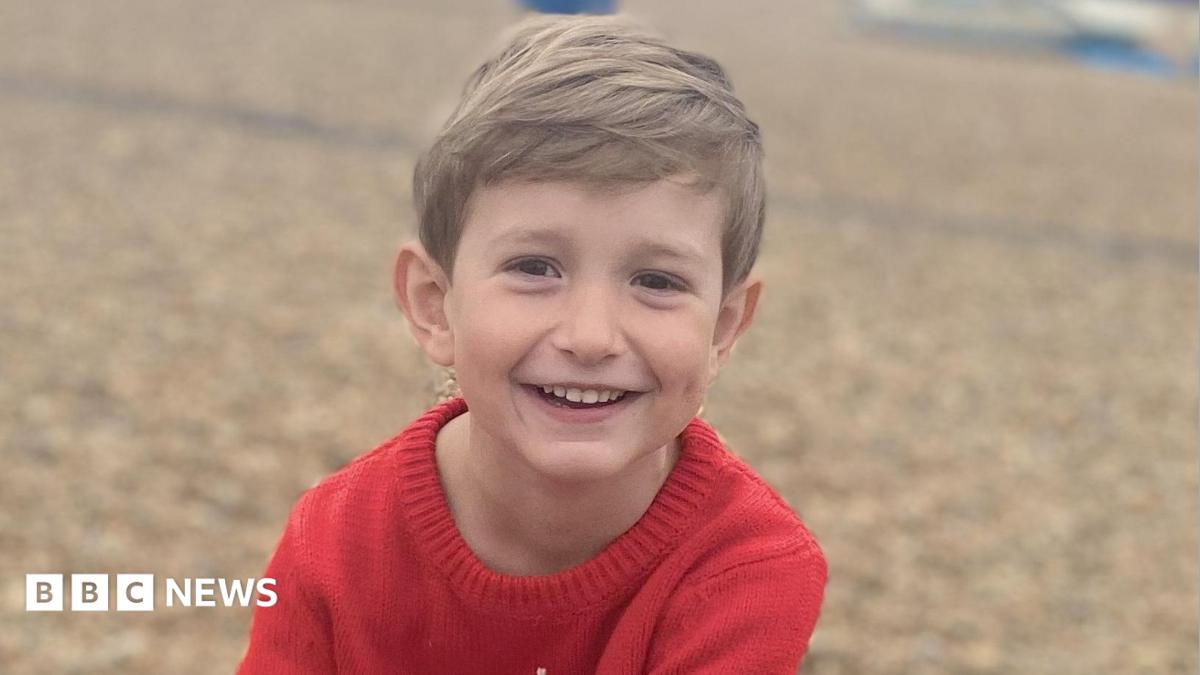 Benedict, a young boy with blond hair, is on a pebbly beach. He is wearing a red knitted top and is smiling to the camera.