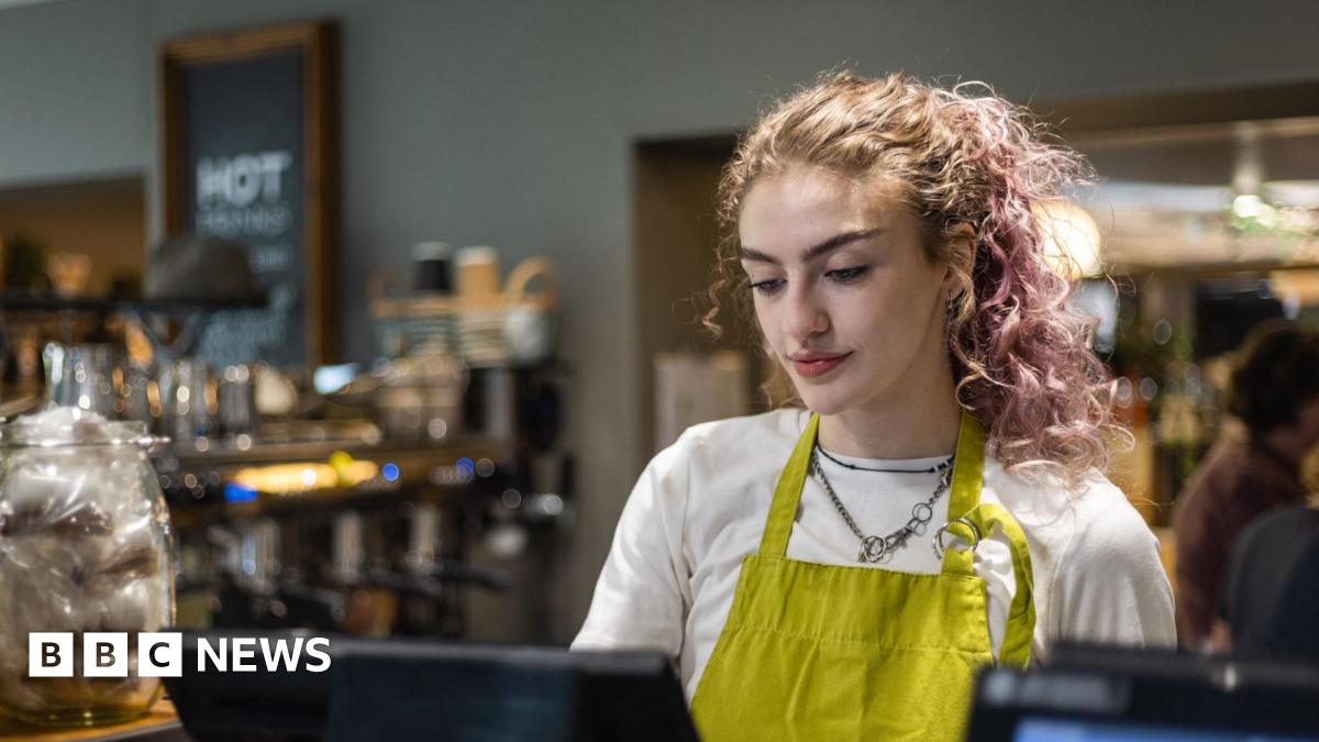 A young woman wearing a green apron looks at a till in a cafe.