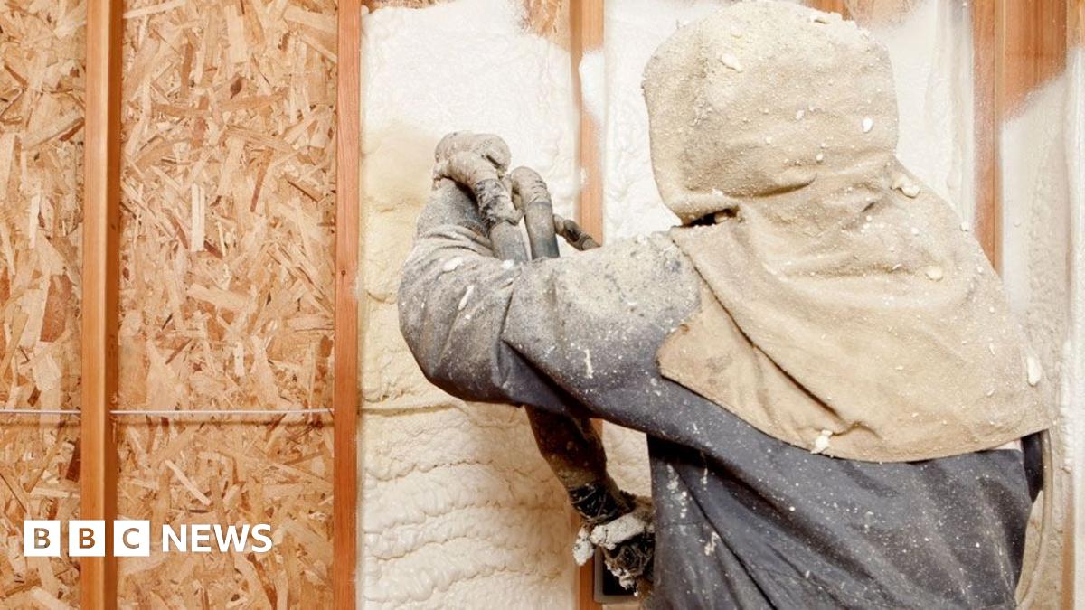 A technician in a protective blue and cream suit uses a spray gun to spray foam against a wall