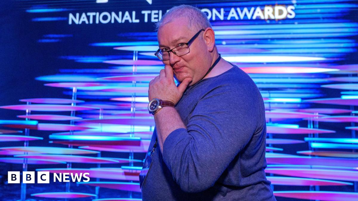 James Kenny is standing sideways in front of a blue and purple promotional screen at the National Television Awards, with the NTA branding behind him. He is a white man with short grey, thinning hair who is smiling and holding his hand up to his chin i...
