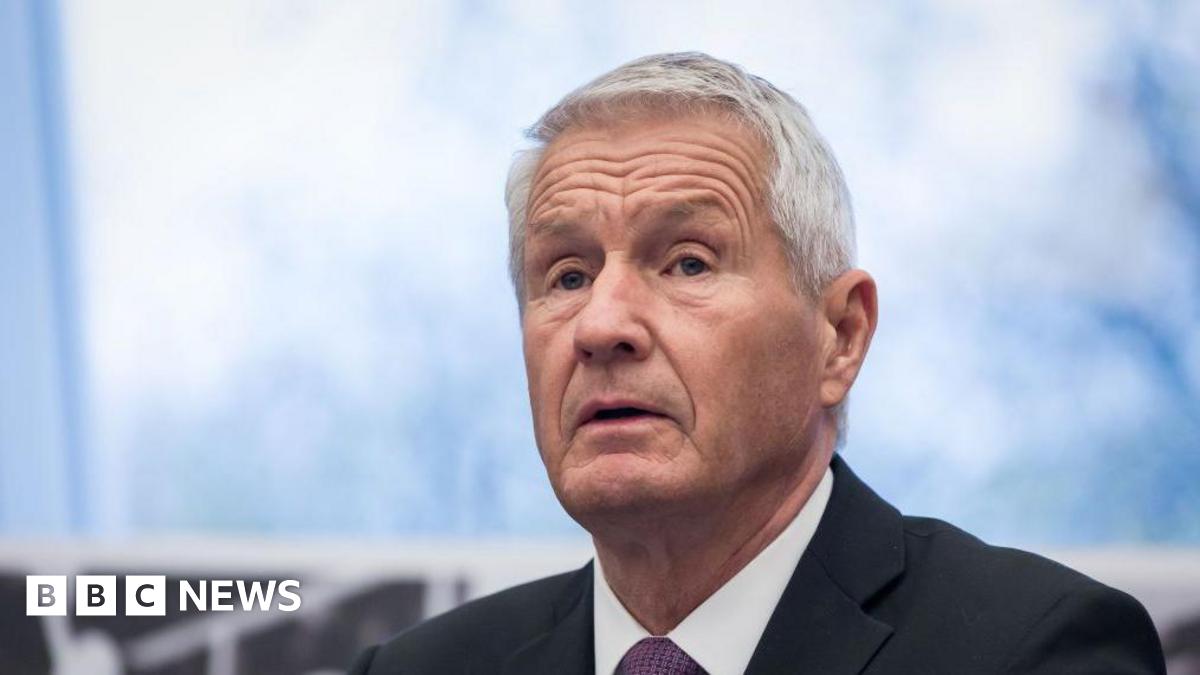 Former Norwegian Prime Minister Thorbjørn Jagland dressed in a black suit jacket, white shirt and a purple tie, he looks off to the left, his mouth tilted down. His hair is grey and skin tanned. The background is blurred. 