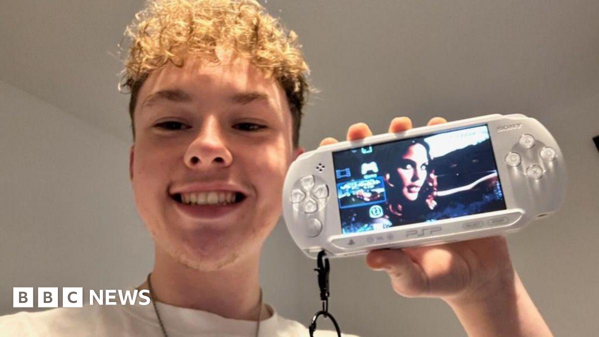 Kyle is indoors holding up a white PlayStation Portable (PSP) console with a keychain attached.