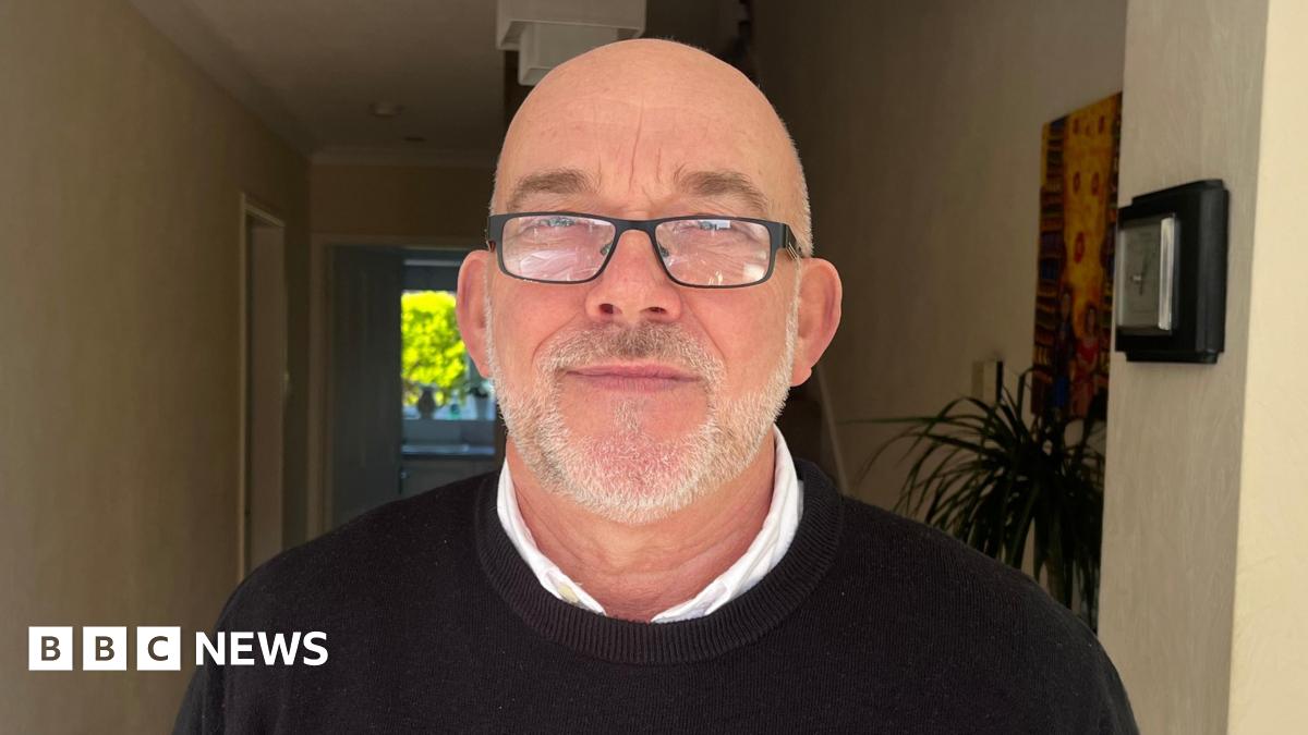 A man with balding grey hair and a beard and wearing a white shirt and dark coloured jumper is smiling as he stands inside the hallway of a house.