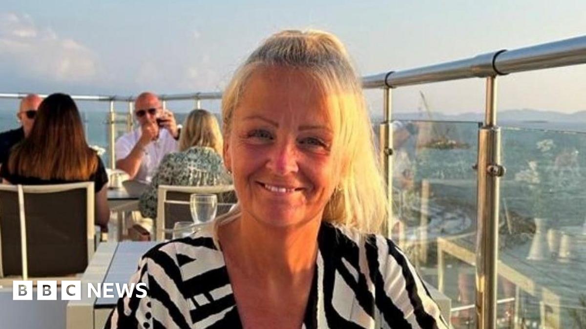 Anita Rose in a black and white top sat looking towards the camera with other people behind, next to a glass balcony. She is smiling with blonde hair