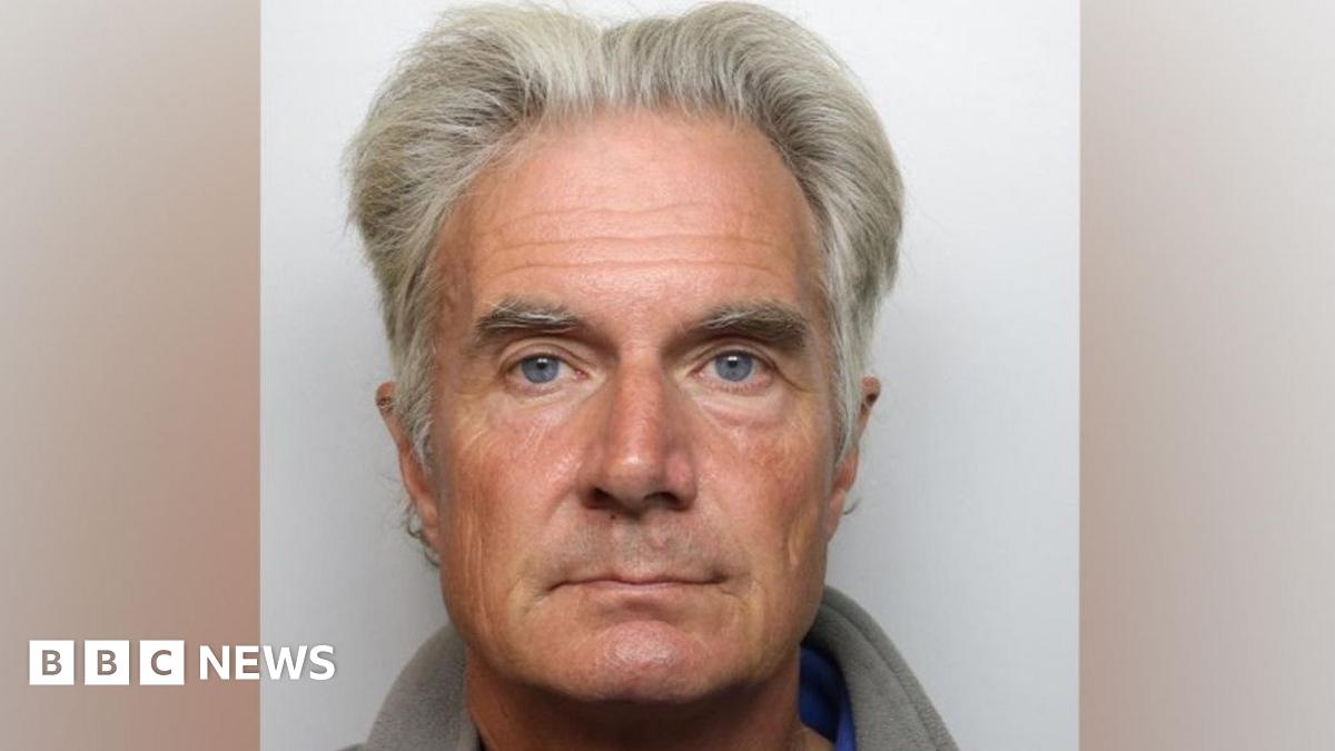 A close up of a mug shot showing an older man with grey hair and eyebrows. He has blue eyes.