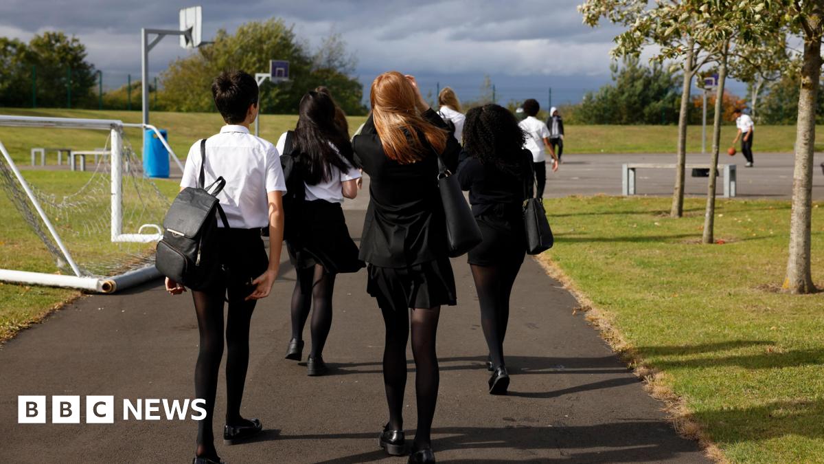 A group of secondary school students walk together across a school ground. They have their backs to the camera but they are wearing black uniforms with a white shirt. They are walking towards a tarmac playground surrounded by grassed areas, trees and s...