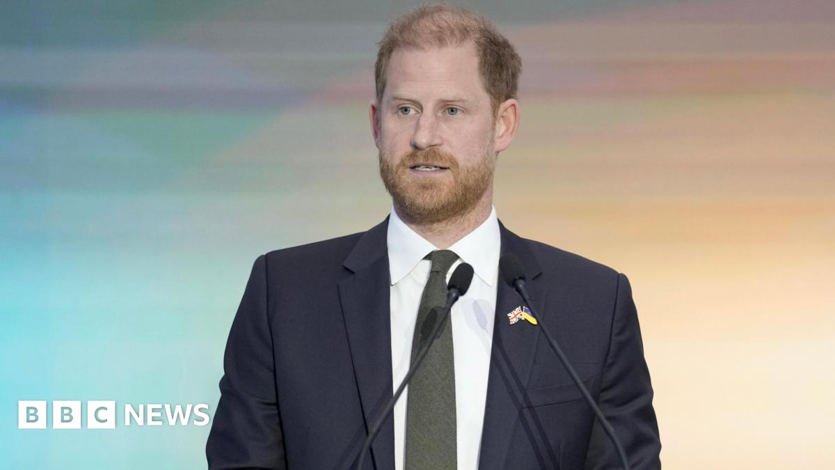 Prince Harry - a man in a dark business suit, white shirt and olive green tie, stands in front of two small microphones as he delivers a speech. The background goes from blue to sunrise yellow in a gentle gradient.