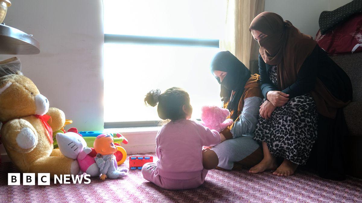 Photo of a hotel room with three people - a women and older child with their faces covered, and a younger girl with a back to the camera sitting on a patterned pink carpet