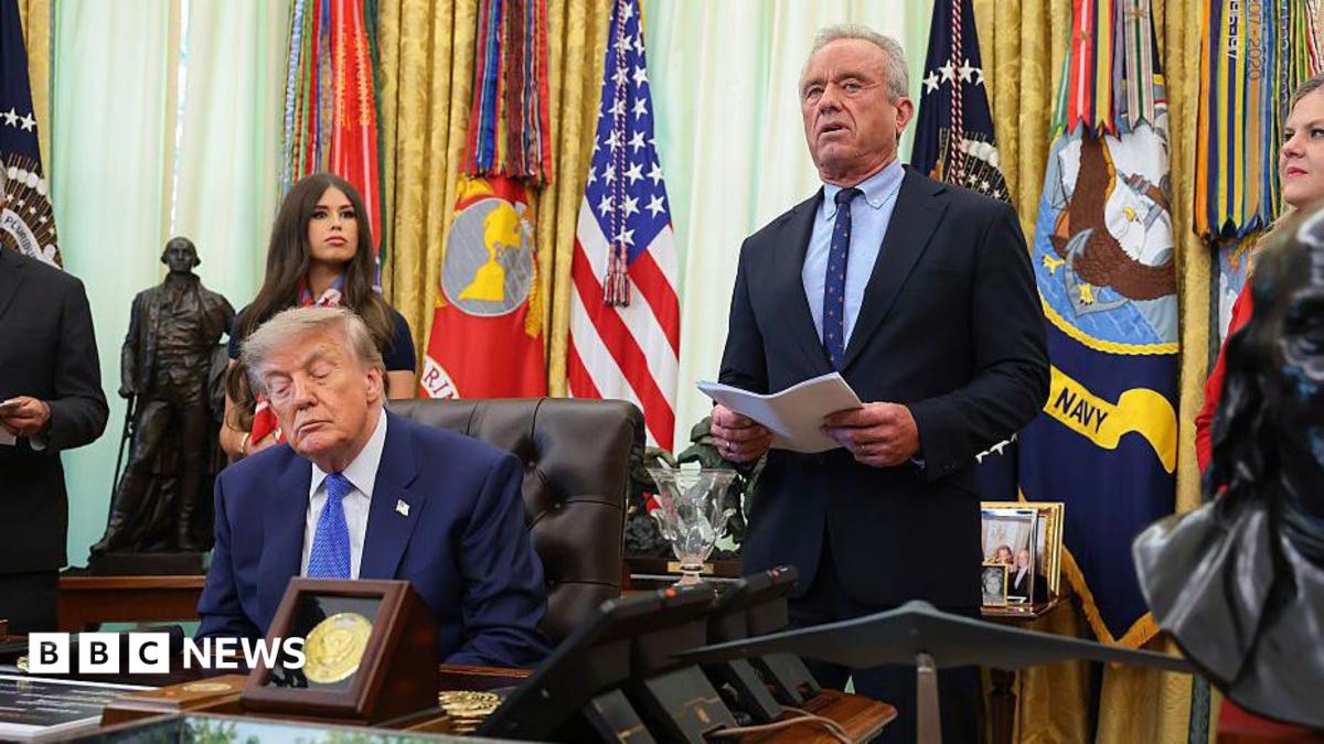A man at a podium speaking while another man sits at a desk in a formal setting with flags.
