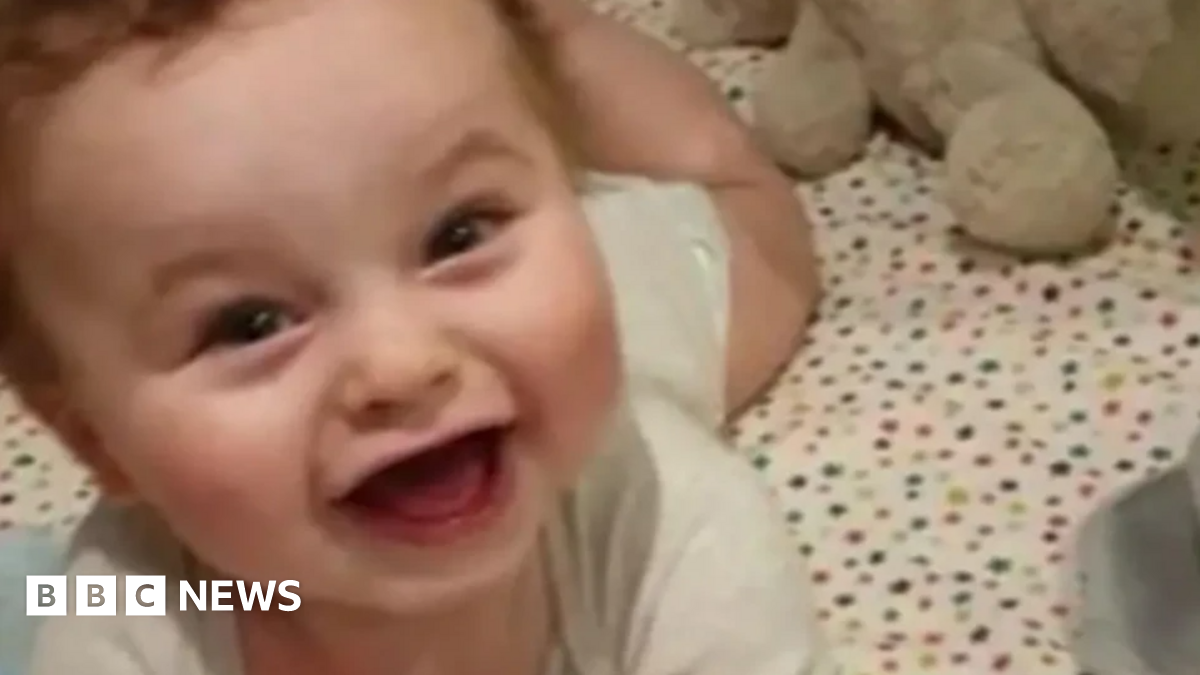 Baby Preston Davey smiles while dressed in a white vest. He appears to be lying in a cot.
